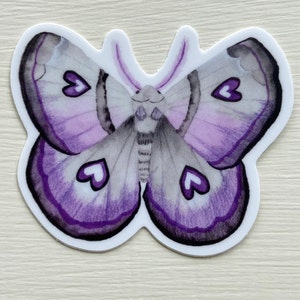 May include: A purple and gray butterfly sticker with a watercolor design. The butterfly has a black outline and white hearts on its wings.