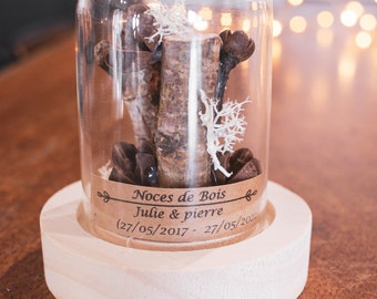 May include: A glass cloche with a wooden base, containing small branches, pinecones, and white dried flowers. The label reads "Noces de Bois Julie & Pierre (27/05/2017 - 27/05/202".