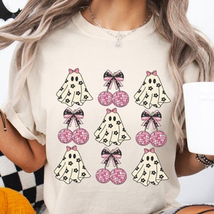 May include: A light beige t-shirt with a pattern of pink bows, white ghosts with black floral patterns, and pink disco balls. The ghosts are wearing pink bows.