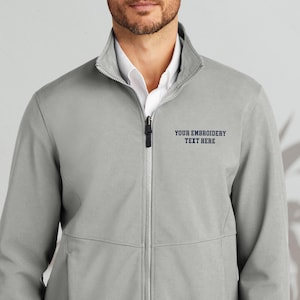 May include: A light gray zip-up jacket with a stand-up collar. The jacket has a white shirt underneath and the text "YOUR EMBROIDERY TEXT HERE" in navy blue. The jacket has two front pockets and a zipper closure.