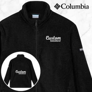 K&ouml;nnte beinhalten: Schwarze Fleecejacke mit durchgehendem Rei&szlig;verschluss und hohem Kragen. Das Columbia-Logo befindet sich oben rechts, und die Worte "Custom Embroidered" sind in wei&szlig;er Schrift geschrieben. Ein kreisf&ouml;rmiger Einsatz zeigt die R&uuml;ckseite der Jacke.