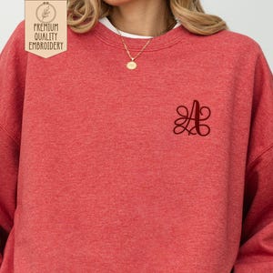 May include: A red sweatshirt with a dark red embroidered letter "A" on the chest. The sweatshirt has a crew neck and long sleeves. The upper left corner of the image has a logo that says "PREMIUM QUALITY EMBROIDERY".