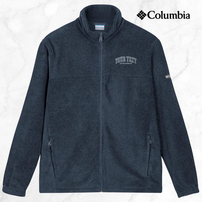 K&ouml;nnte beinhalten: Marineblaue Fleecejacke mit durchgehendem Rei&szlig;verschluss und hohem Kragen. Die Jacke zeigt das Columbia-Logo und den gestickten Text "YOUR TEXT" auf der Brust. Sie hat zwei Seitentaschen und ein kleines Logo am &Auml;rmel.