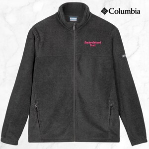 K&ouml;nnte beinhalten: Dunkelgraue Fleecejacke mit durchgehendem Rei&szlig;verschluss und hohem Kragen. Die Jacke hat zwei Rei&szlig;verschlusstaschen und das Columbia-Logo oben rechts. Auf der linken Brust ist rosa gestickter Text zu sehen.