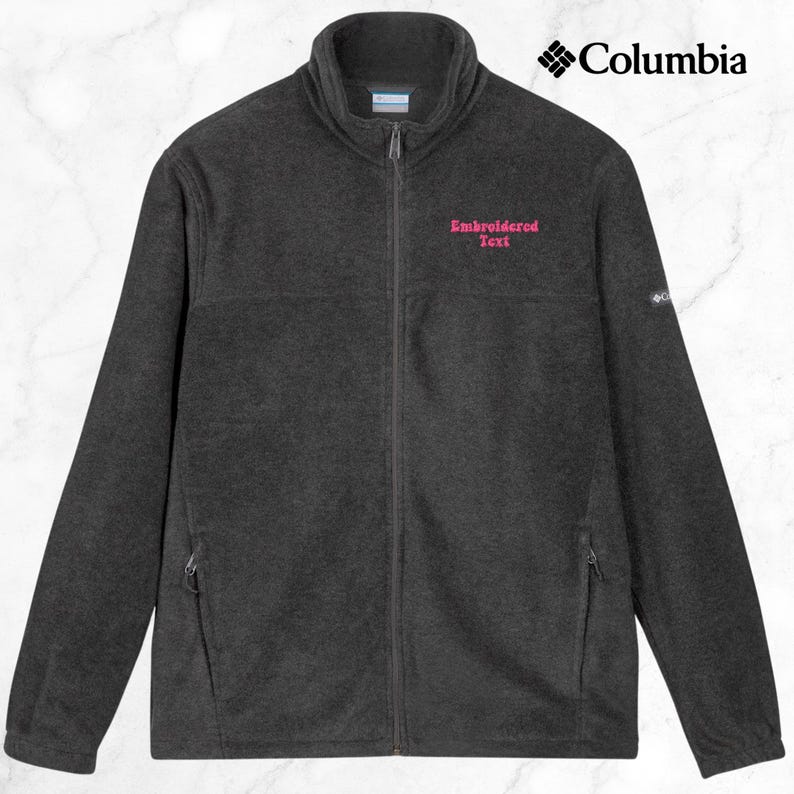 K&ouml;nnte beinhalten: Dunkelgraue Fleecejacke mit durchgehendem Rei&szlig;verschluss und hohem Kragen. Die Jacke hat zwei Rei&szlig;verschlusstaschen und das Columbia-Logo oben rechts. Auf der linken Brust ist rosa gestickter Text zu sehen.