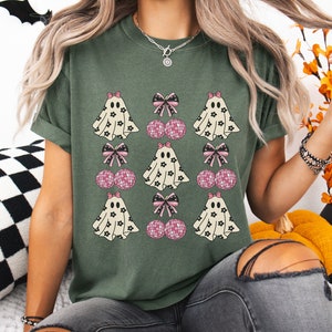 Puede incluir: Una camiseta verde con un patr&oacute;n repetido de fantasmas y cerezas rosas con lazos. Los fantasmas son blancos con dise&ntilde;os de flores negras y las cerezas son rosas con brillo.