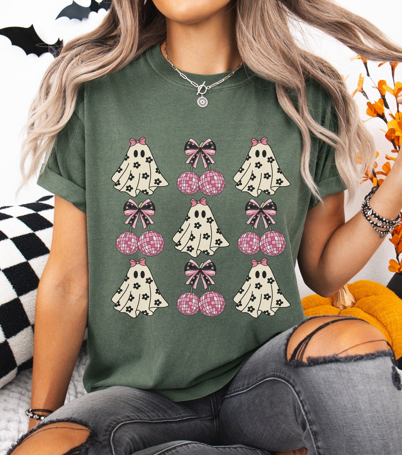 Puede incluir: Una camiseta verde con un patr&oacute;n repetido de fantasmas y cerezas rosas con lazos. Los fantasmas son blancos con dise&ntilde;os de flores negras y las cerezas son rosas con brillo.