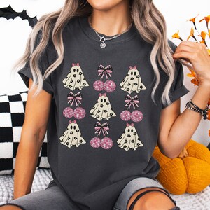 Puede incluir: Una camiseta gris oscuro con un estampado gr&aacute;fico de fantasmas blancos con patrones florales negros y lazos rosas. Los fantasmas est&aacute;n emparejados con lazos rosas y cerezas de bola de discoteca.