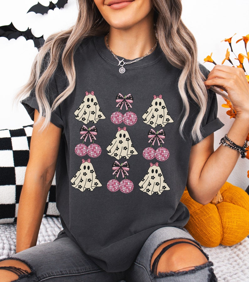 Puede incluir: Una camiseta gris oscuro con un estampado gr&aacute;fico de fantasmas blancos con patrones florales negros y lazos rosas. Los fantasmas est&aacute;n emparejados con lazos rosas y cerezas de bola de discoteca.