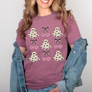 Puede incluir: Una camiseta malva con un estampado gr&aacute;fico de fantasmas con lazos rosas y cerezas de lunares rosas y negros.