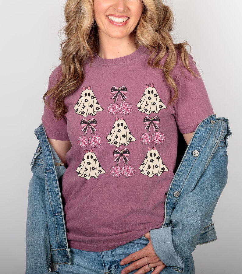Puede incluir: Una camiseta malva con un estampado gr&aacute;fico de fantasmas con lazos rosas y cerezas de lunares rosas y negros.