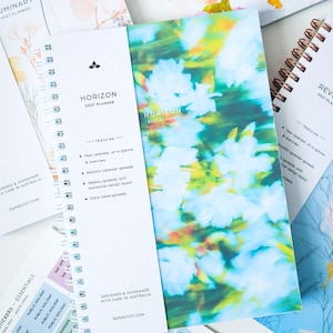 以下が含まれることがあります： スパイラル綴じのHorizon 2027プランナー。Horizon 2027 Planner、features、designed and handmade with care in Australiaという文字が印刷された白い紙の帯が付いています。表紙は青と緑の鮮やかなぼかし花柄です。