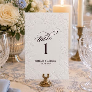 May include: A white rectangular table number card featuring an embossed floral border, black script text reading Table 1, and the names PHILLIP & ASHLEY with the date 09.13.2026 printed in black, held upright by a small gold-toned metal stand.