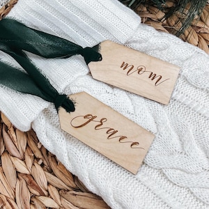 May include: Two rectangular light-colored wooden tags with dark green ribbon ties. Each tag features custom text engraved in a cursive script. One tag displays the word mom and the other tag displays the word grace. The tags are placed on a white knit fabric.