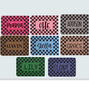 May include: Rectangular personalized nameplates with a checkered pattern in various colors, including pink, black, brown, blue, green, and purple. Each plate features a name or word in a contrasting color, such as "mama", "Gavin", and "Claire".