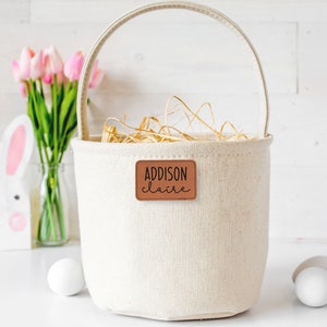 May include: A white canvas Easter basket with a brown leather tag that says "ADDISON Claire". The basket is filled with straw and there are white Easter eggs on the table.