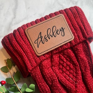 May include: Red knitted stocking with a brown leather patch that says "Ashley" in black script.