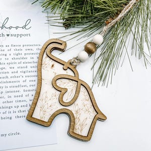 Puede incluir: Adorno de madera con forma de elefante con un dise&ntilde;o floral, ensartado con un cord&oacute;n de yute y cuentas de madera. El adorno est&aacute; hecho de madera de color claro con un contorno de madera m&aacute;s oscuro. Una ramita de pino est&aacute; en el fondo.