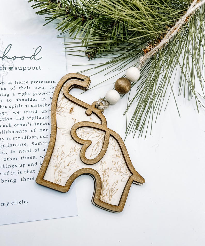 Puede incluir: Adorno de madera con forma de elefante con un dise&ntilde;o floral, ensartado con un cord&oacute;n de yute y cuentas de madera. El adorno est&aacute; hecho de madera de color claro con un contorno de madera m&aacute;s oscuro. Una ramita de pino est&aacute; en el fondo.
