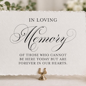 May include: A rectangular white card with a deckled edge featuring black printed text that reads IN LOVING MEMORY OF THOSE WHO CANNOT BE HERE TODAY BUT ARE FOREVER IN OUR HEARTS. The card is held upright by a small gold-toned metal stand.