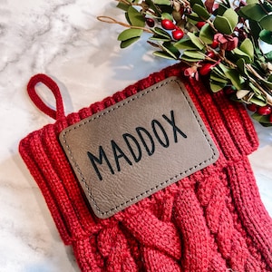 May include: Red knitted Christmas stocking with a brown leather name tag that says "MADDOX".