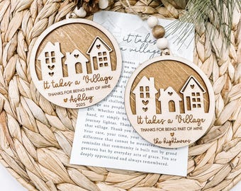 May include: Two round wooden ornaments with a village design and the text "It takes a Village." One ornament is personalized with a name and the year 2025. The ornaments are on a woven background.
