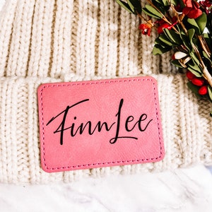 May include: Cream-colored knit beanie with a pink rectangular patch. The patch is stitched to the beanie and features the name "Finn Lee" in black cursive script. The beanie is made of a thick, ribbed knit material.