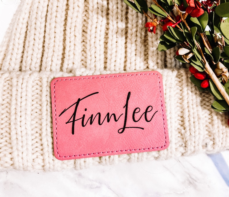 May include: Cream-colored knit beanie with a pink rectangular patch. The patch is stitched to the beanie and features the name "Finn Lee" in black cursive script. The beanie is made of a thick, ribbed knit material.