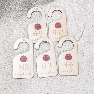 May include: Set of five wooden closet dividers for baby clothes. Each divider has a hanger top and features a pink seashell design. The dividers are labeled with age ranges: NB newborn, 0-3 months, 3-6 months, 6-9 months, and 9-12 months.