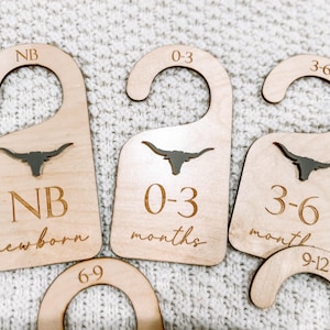 Puede incluir: Cuatro perchas de madera con un recorte de cabeza de toro longhorn. Las perchas están etiquetadas con diferentes tamaños: NB (recién nacido), 0-3 meses, 3-6 meses y 6-9 meses.