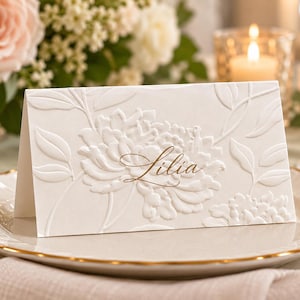 May include: A white folded place card featuring an embossed floral pattern and the name Lilia written in elegant brown script. The card is made of heavy cardstock with a textured surface, displaying a detailed relief of blooming flowers and leaves.