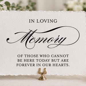 May include: A rectangular white card with a deckled edge featuring black printed text that reads IN LOVING MEMORY OF THOSE WHO CANNOT BE HERE TODAY BUT ARE FOREVER IN OUR HEARTS. The card is held upright by a small gold-toned metal stand.
