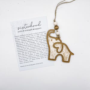Puede incluir: Adorno de madera con forma de elefante con un dise&ntilde;o floral, colgado de una cuerda con cuentas de madera. Una tarjeta con la palabra "sisterhood" y texto sobre la unidad, la fuerza y el apoyo est&aacute; al lado del adorno.