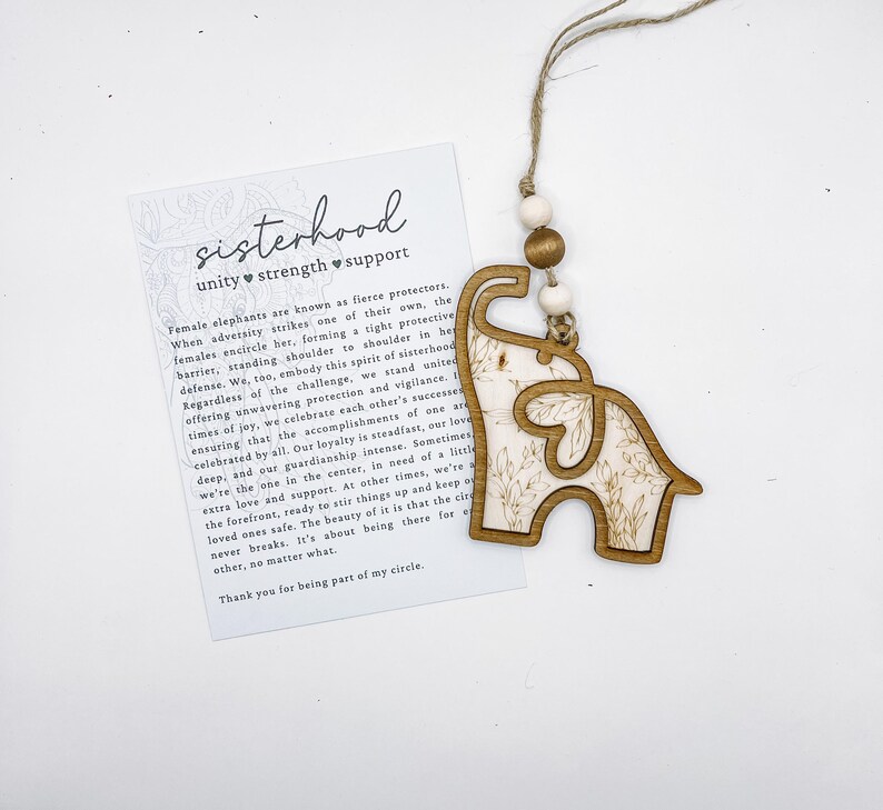 Puede incluir: Adorno de madera con forma de elefante con un dise&ntilde;o floral, colgado de una cuerda con cuentas de madera. Una tarjeta con la palabra "sisterhood" y texto sobre la unidad, la fuerza y el apoyo est&aacute; al lado del adorno.