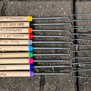 May include: A collection of s'mores roasting sticks with wooden handles and metal prongs. Each handle has a different color accent and is engraved with phrases like "Spread S'more Love" and names like "Emery."