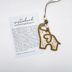Puede incluir: Adorno de madera con forma de elefante y dise&ntilde;o floral, colgado de una cuerda. El adorno se muestra con una tarjeta con la palabra "sisterhood" y un mensaje sobre unidad, fuerza y apoyo.