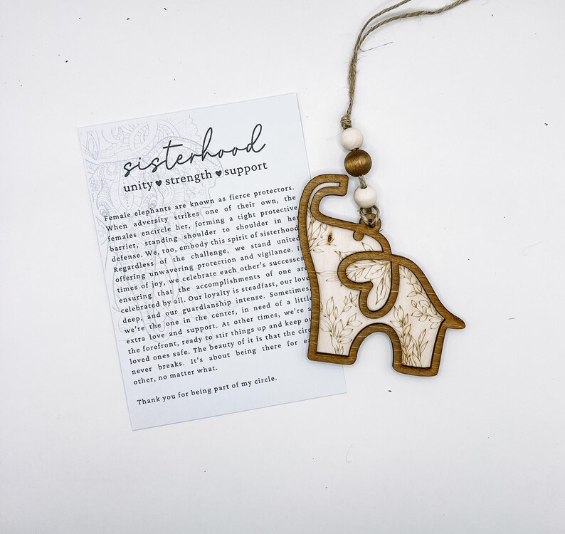Puede incluir: Adorno de madera con forma de elefante y dise&ntilde;o floral, colgado de una cuerda. El adorno se muestra con una tarjeta con la palabra "sisterhood" y un mensaje sobre unidad, fuerza y apoyo.