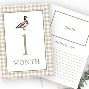 May include: A printable baby milestone card with a gingham background. The card features a watercolor illustration of a duck and the number 1 with the text "1 month". The card also has a space for a photo and a section for milestones.