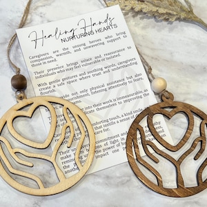 May include: Two wooden ornaments with a heart and hands design. The ornaments are brown and natural wood. The text on the card reads "Healing Hands Nurturing Hearts".