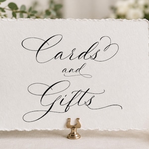 May include: A rectangular white card with a deckled edge featuring the black calligraphic text Cards and Gifts. The card is held upright by a small gold-toned metal stand. The design is centered on the textured paper surface.