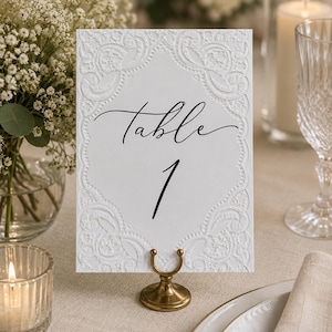 May include: A white rectangular table number card featuring an embossed lace-like border and the black script text Table 1, held upright by a small, circular, antique brass-colored metal stand. The card has a textured surface with intricate patterns.