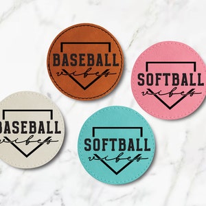 May include: Four round leather coasters with black baseball and softball designs. The coasters are brown, pink, beige, and turquoise.