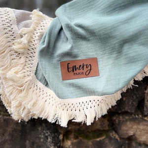 May include: A light blue baby blanket with a white crocheted trim and a brown leather tag that says "Emery Paige".