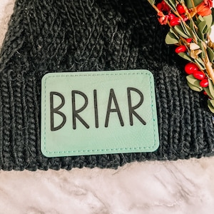 May include: A black knit beanie with a rectangular mint green patch that reads "BRIAR" in black letters. The beanie is made of a thick, textured yarn. Red berries and greenery are visible in the background.