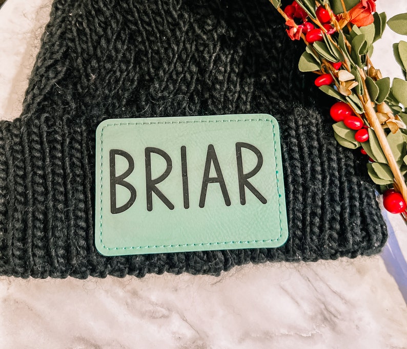 May include: A black knit beanie with a rectangular mint green patch that reads "BRIAR" in black letters. The beanie is made of a thick, textured yarn. Red berries and greenery are visible in the background.