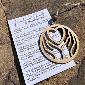 May include: Wooden ornament featuring a heart and two hands, representing healing and nurturing hearts. The ornament is beige and has a string for hanging. The text on the card reads: "Healing Hands Nurturing Hearts Caregivers are the unsung heroes who bring compassion, warmth, and unwavering support to those in need. Their presence brings solace and reassurance to individuals who may feel vulnerable or isolated. With gentle gestures and soothing creates a safe space where trust can flourish. They provide not only physical nourishment, emotional nourishment, hopes, and dreams. The love they pour into the lives of others. They selflessly dedicate their lives to others. Caregivers offer a comforting and reassuring presence that instills worth in those they care for. Their unwavering commitment to make them beacons of light in reminding us of the power of human connection. The profound impact one person can have on another's life."