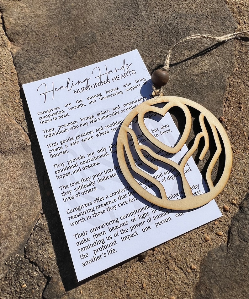 May include: Wooden ornament featuring a heart and two hands, representing healing and nurturing hearts. The ornament is beige and has a string for hanging. The text on the card reads: "Healing Hands Nurturing Hearts Caregivers are the unsung heroes who bring compassion, warmth, and unwavering support to those in need. Their presence brings solace and reassurance to individuals who may feel vulnerable or isolated. With gentle gestures and soothing creates a safe space where trust can flourish. They provide not only physical nourishment, emotional nourishment, hopes, and dreams. The love they pour into the lives of others. They selflessly dedicate their lives to others. Caregivers offer a comforting and reassuring presence that instills worth in those they care for. Their unwavering commitment to make them beacons of light in reminding us of the power of human connection. The profound impact one person can have on another's life."