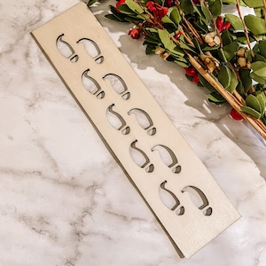 May include: A light-colored wooden Christmas ornament holder with cut-out Santa hat shapes. The holder is rectangular and angled, with nine hat cutouts. Greenery with red berries is in the background.