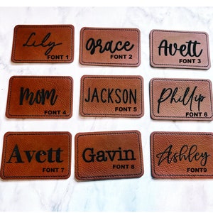 May include: Nine rectangular brown leather patches with black text. The patches display names such as "Lily", "Grace", "Avett", "Mom", "Jackson", "Phillip", "Gavin", and "Ashley". Each patch has a different font style and the text "FONT" followed by a number.