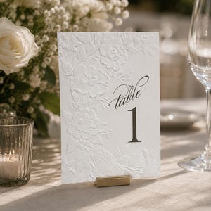 May include: A white rectangular table number card featuring an embossed floral pattern on the left side and the black printed text Table 1 on the right. The card is held upright by a small gold metal stand on a light-colored fabric surface.